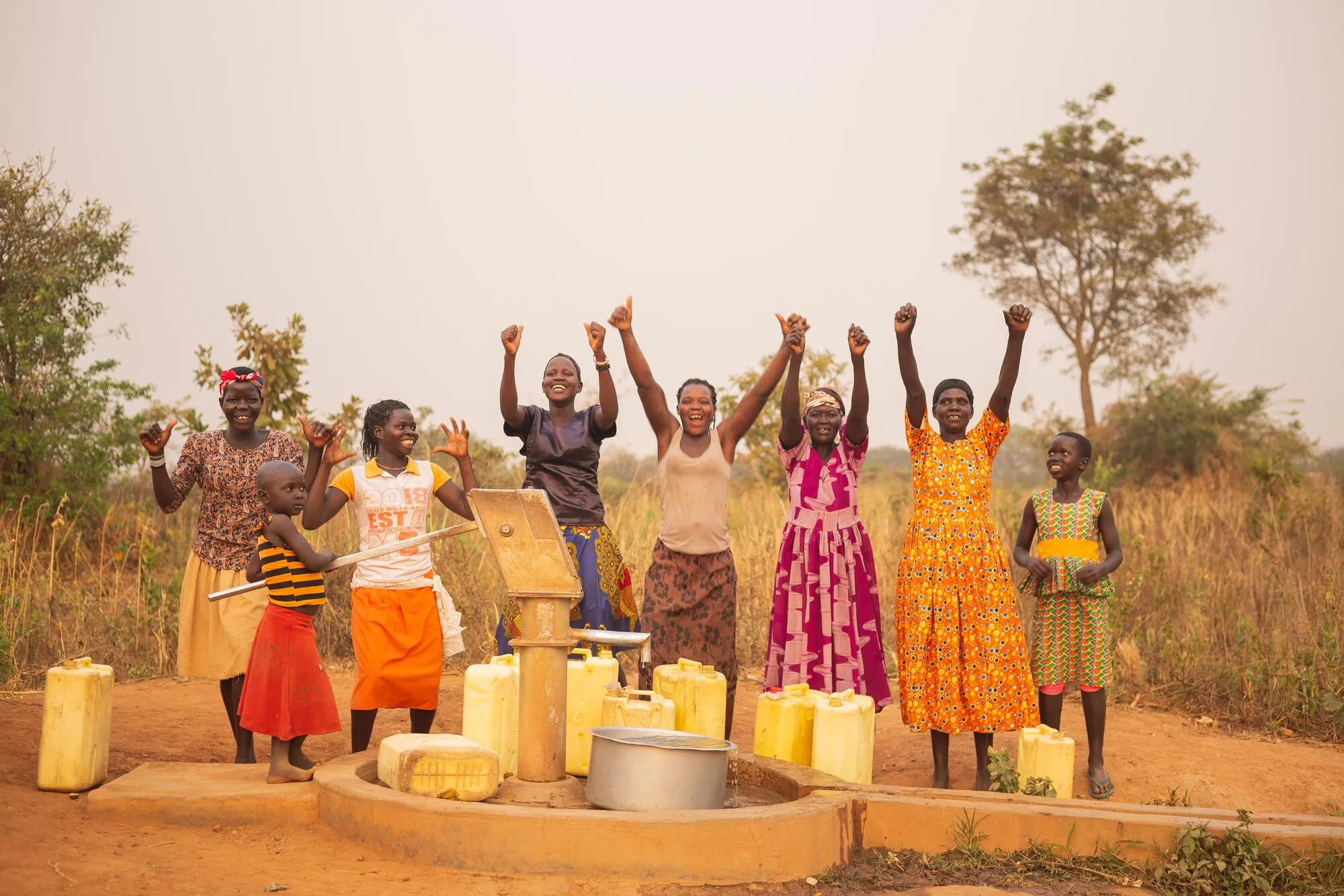 Women and children cheering beside a borehole, with yellow water containers