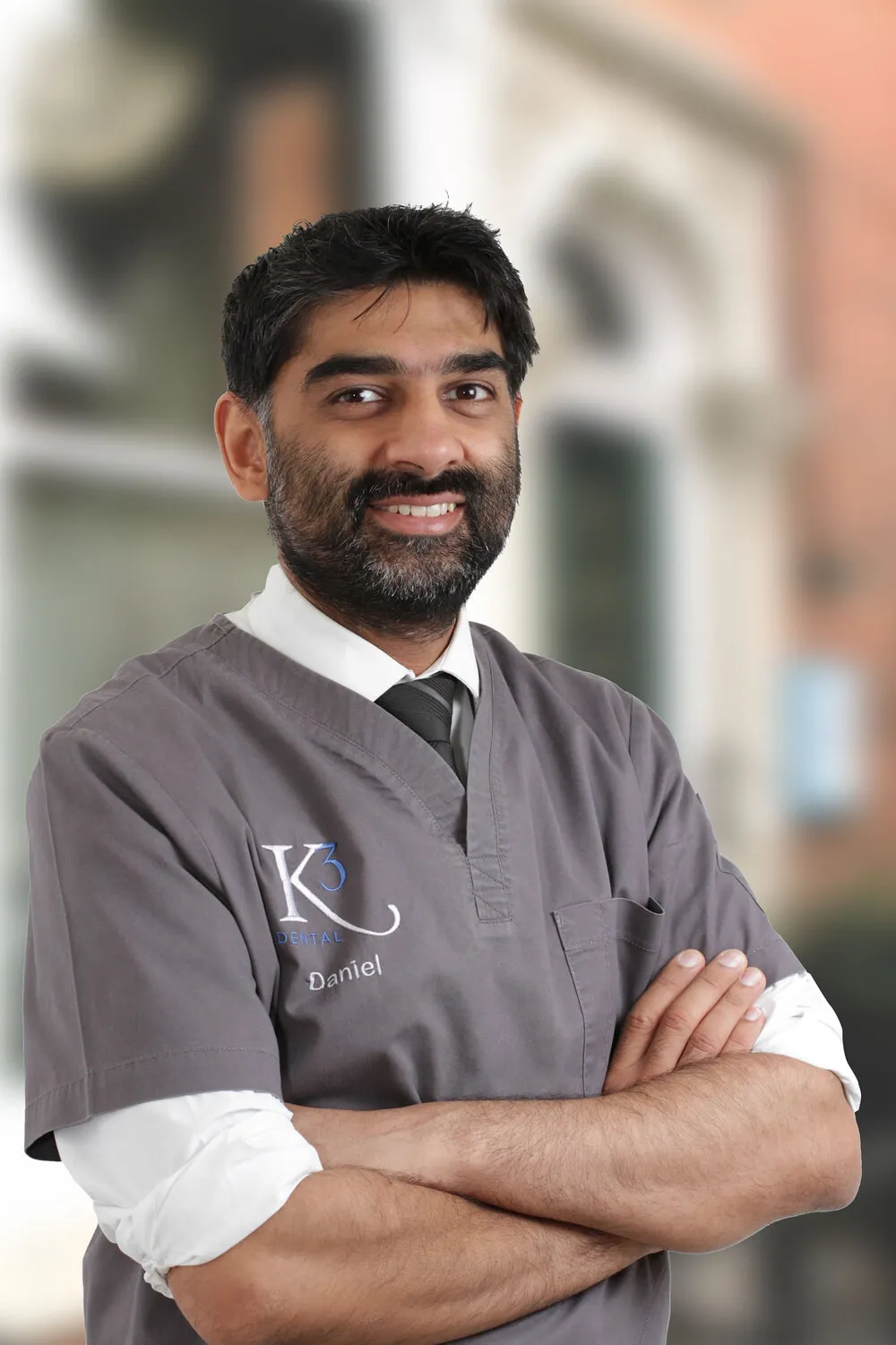 Mr Daniel Saund, arms folded, in K3 scrubs over a shirt and tie, in front of the practice’s windows