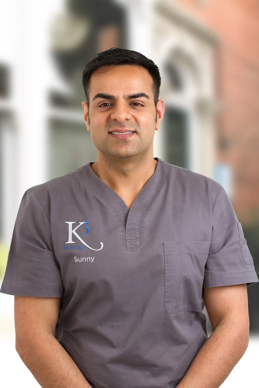 Dr Sunny Kotecha in grey K3 scrubs, in front of the practice’s red brick and white window frames