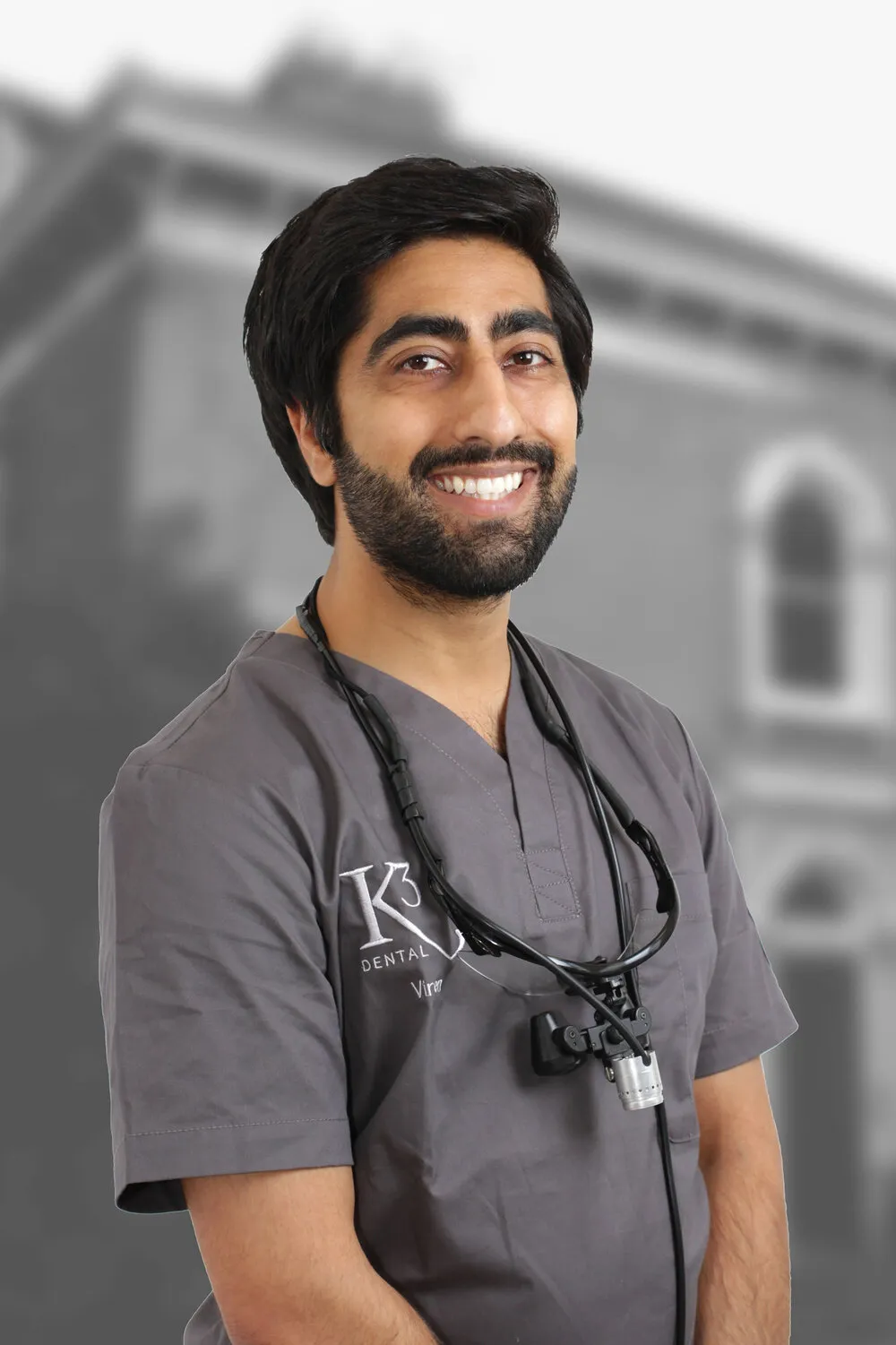 Dr Viren Vithlani in K3 scrubs with loupes round the neck, in front of the building in black and white