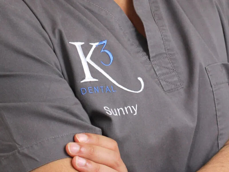 Close-up of grey scrubs with the K3 Dental mark and the name Sunny embroidered in white and blue