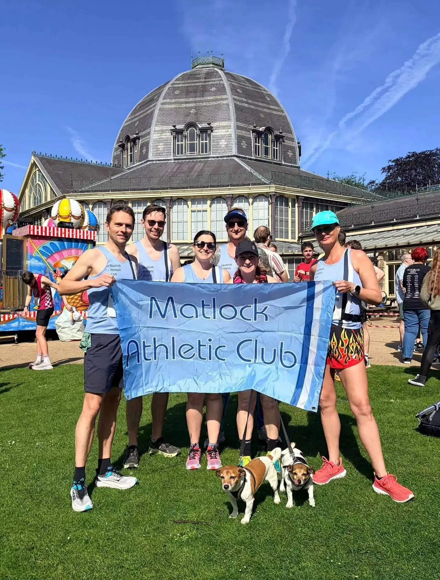 Club members holding a Matlock Athletic Club flag on the grass in front of a Victorian glass pavilion, a small dog at their feet
