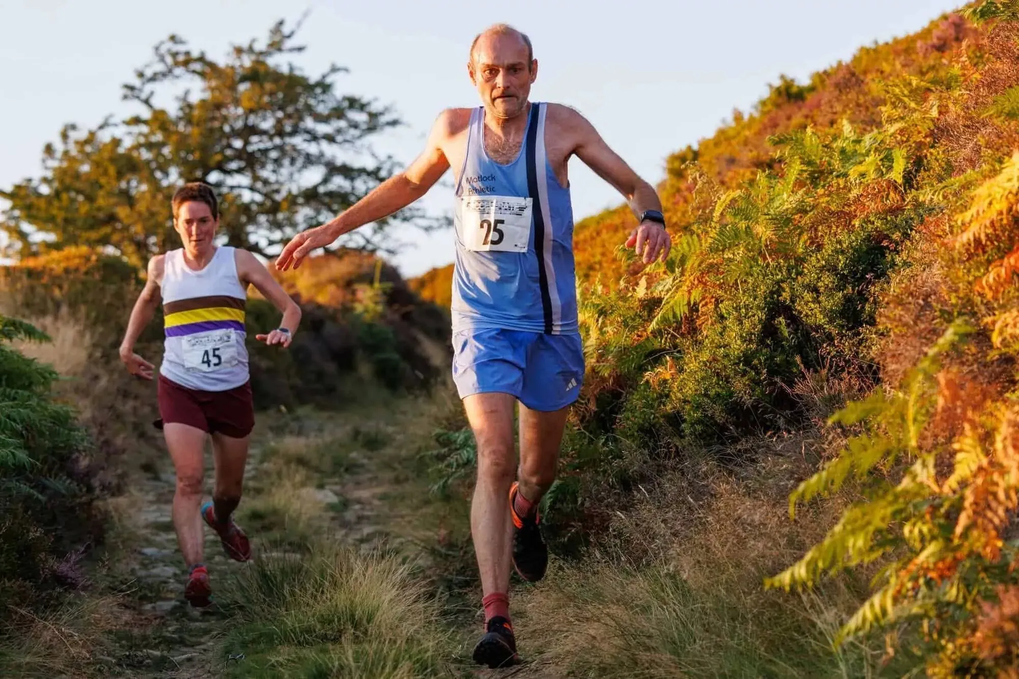 Two runners on a narrow path between golden bracken, a Matlock runner in the club’s pale blue vest in front