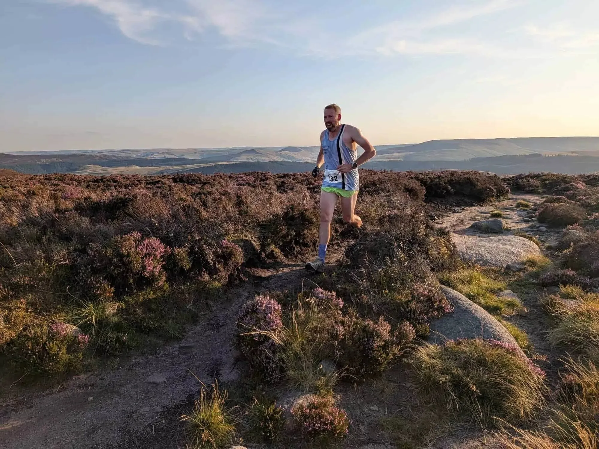 A runner coming down a rocky path through heather on a gritstone edge, the moors beyond in evening light