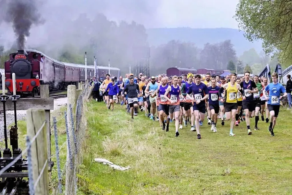 The start of Peak Rail 7: a crowd of runners setting off across a field beside a red steam engine and its carriages, smoke rising