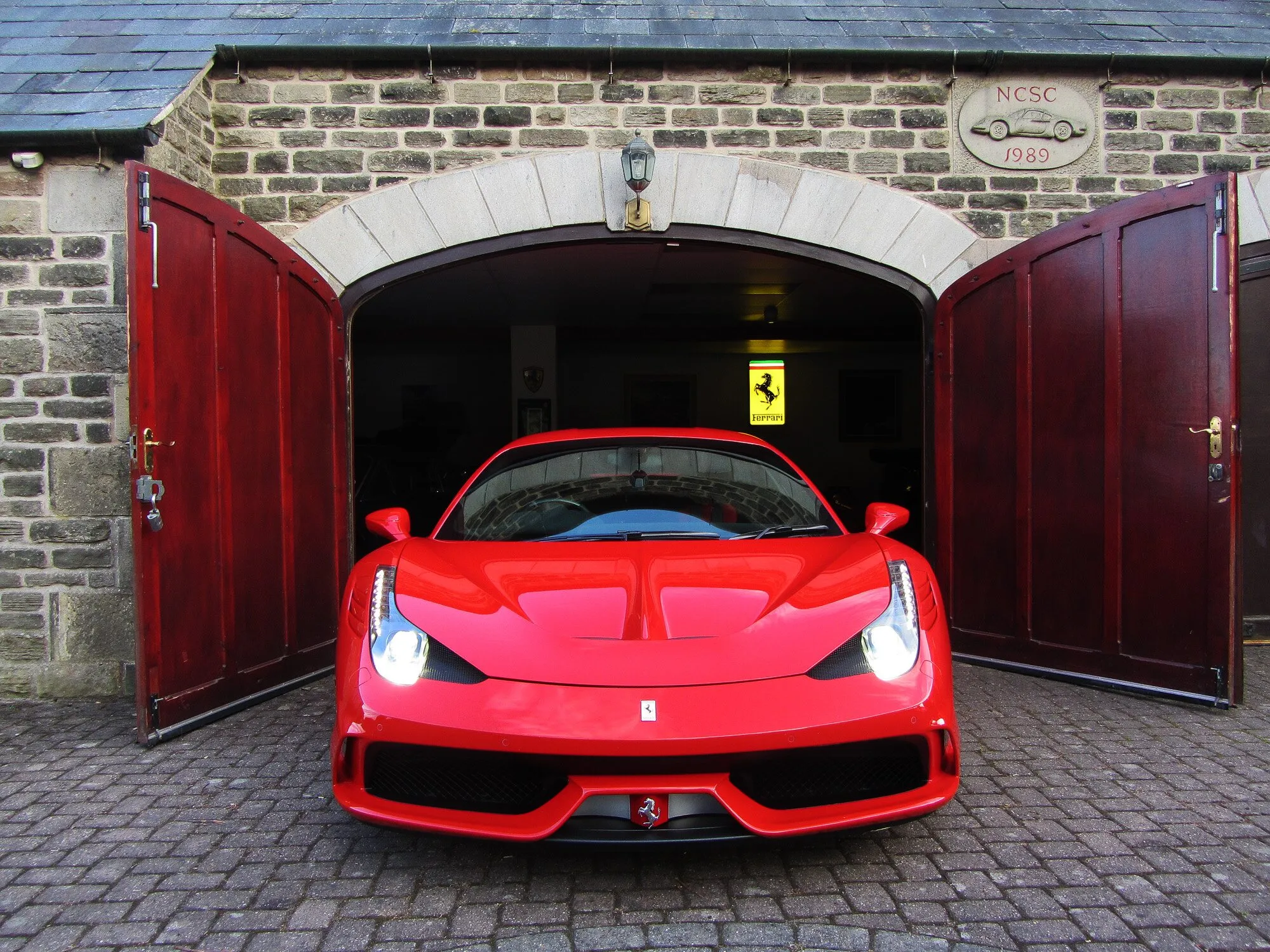 A red Ferrari 458 Speciale in the arched doorway of the stone barn, under the carved NCSC Dino plaque