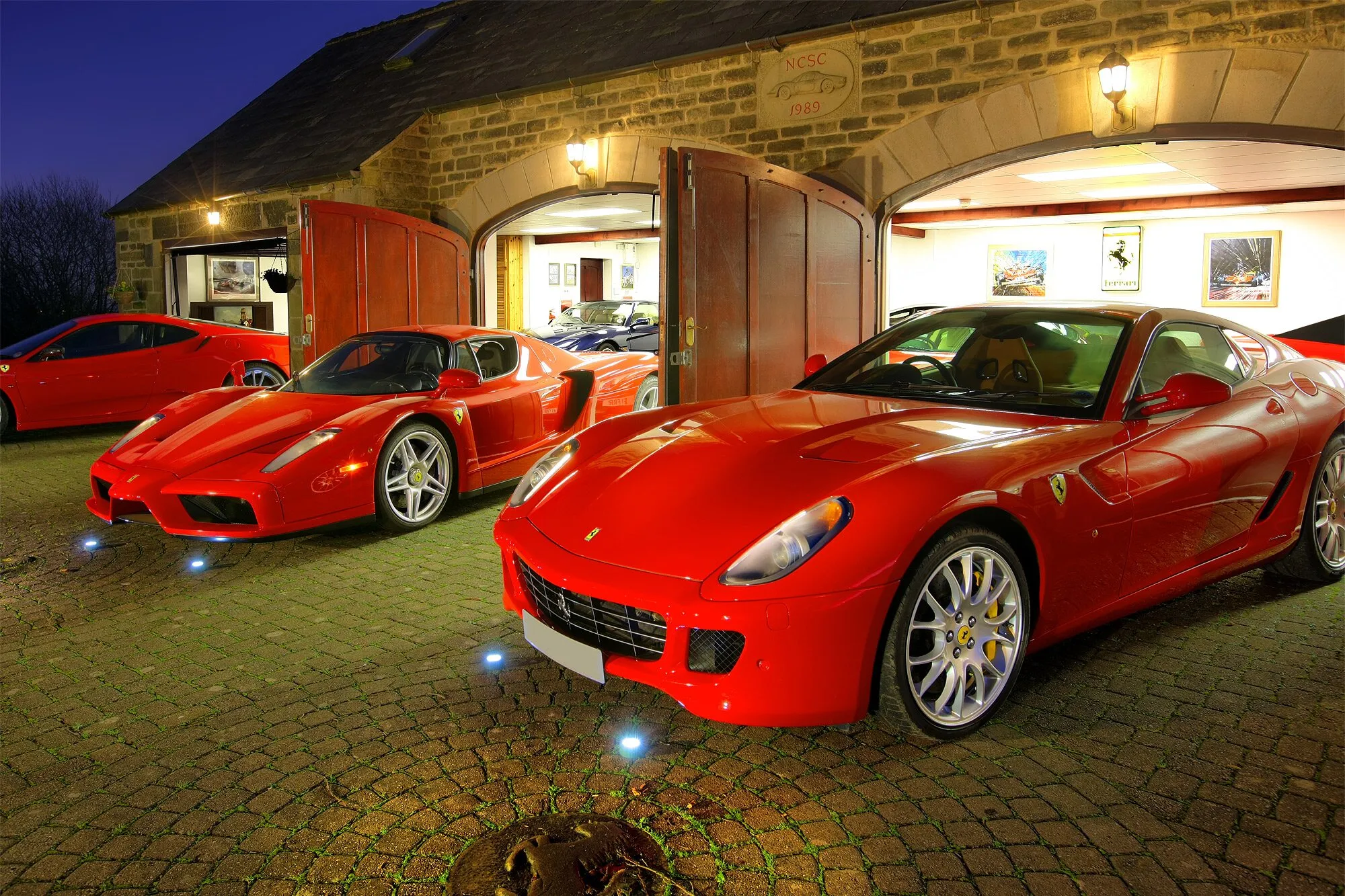 A red Ferrari 599 and an Enzo on the forecourt at night, the barn doors open behind them