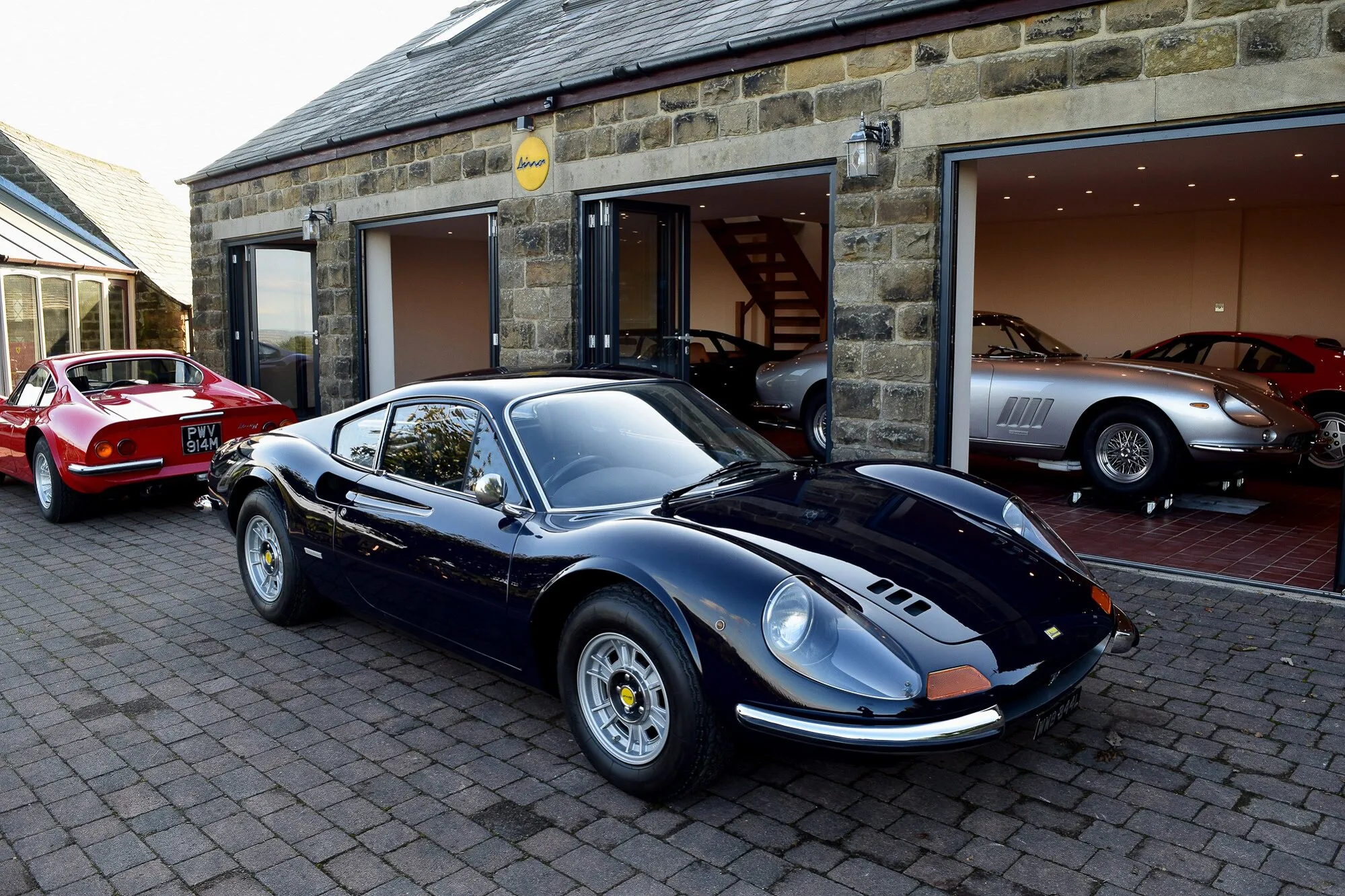 A dark blue Ferrari Dino outside the barns, with more cars inside the open doors