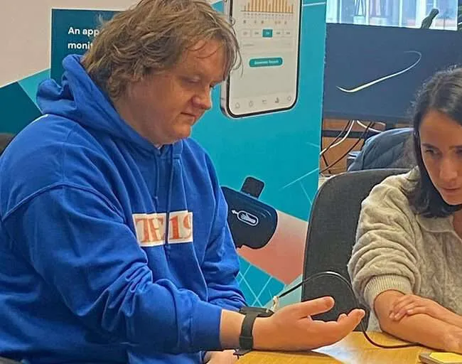 Lewis Capaldi trying the wrist device at the Neupulse office, with the device set up on a laptop alongside and a Neupulse banner stand behind