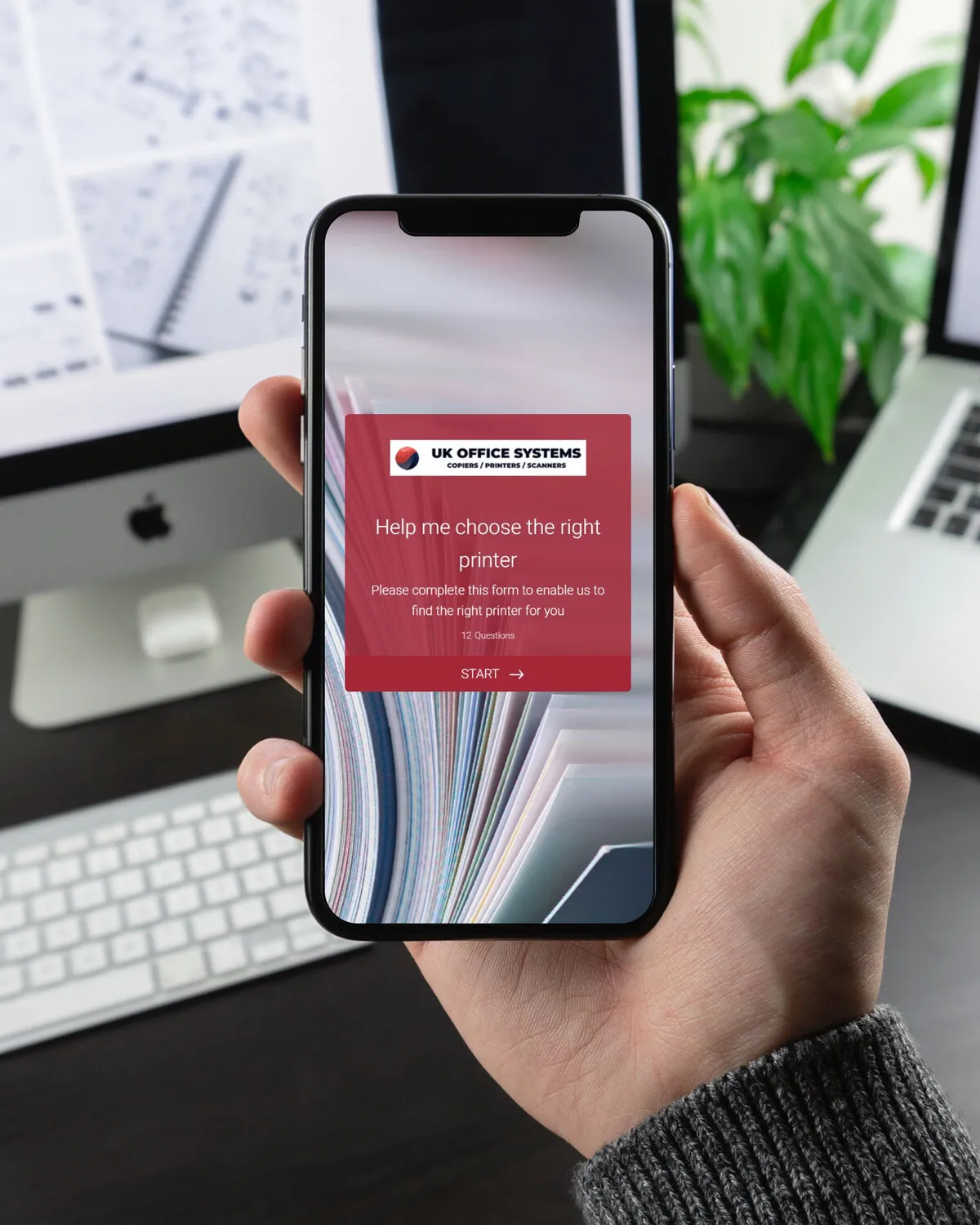Help me choose on a phone held in a hand, in front of a desk with a computer and a laptop: “Help me choose the right printer” in white on UK Office Systems’ red over a close-up of paper, with a Start button