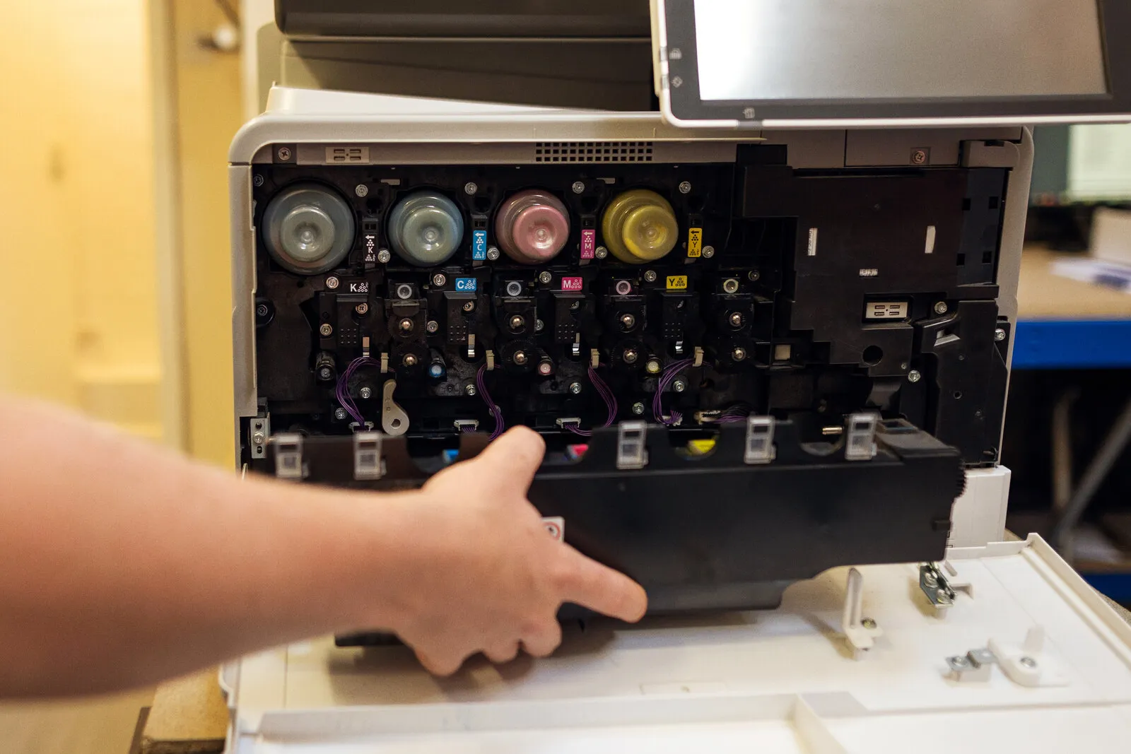 A hand easing out a part inside an open colour copier, below its black, cyan, magenta and yellow toners