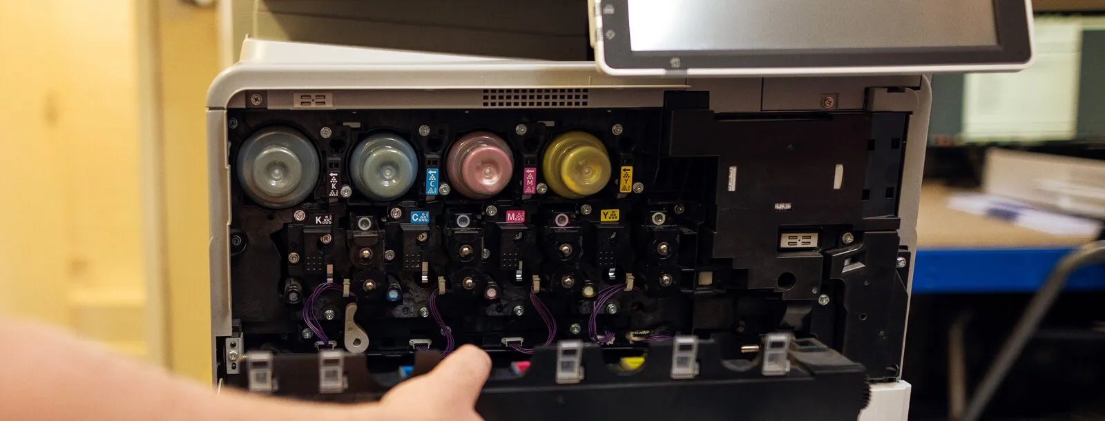 Inside an open Ricoh colour copier: four toner bottles in a row with K, C, M and Y labels for black, cyan, magenta and yellow, and an engineer’s hand easing out the waste toner unit below them