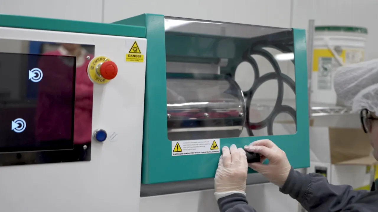 A gloved hand at the door of a seed-treatment machine with a teal frame and a red stop button