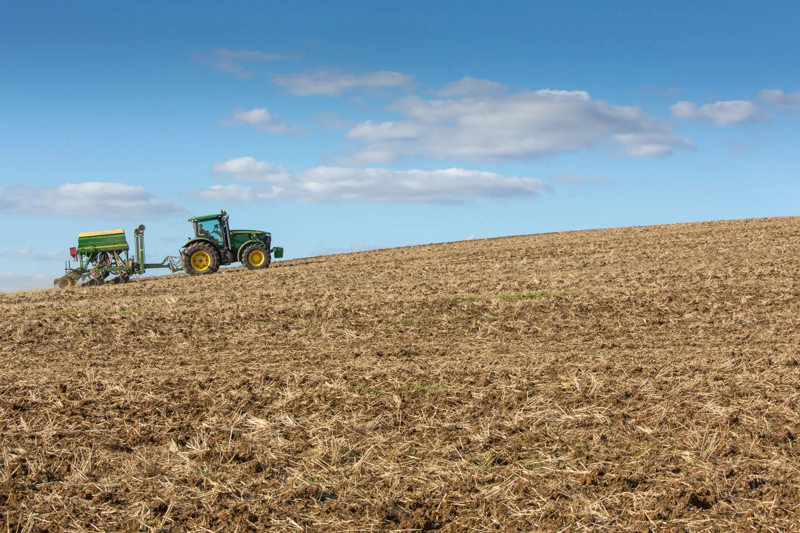 A green tractor drilling seed up a bare brown slope under a blue sky