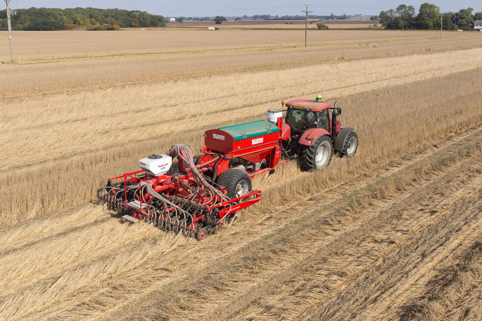 A red tractor and seed drill working across a field of stubble, seen from above