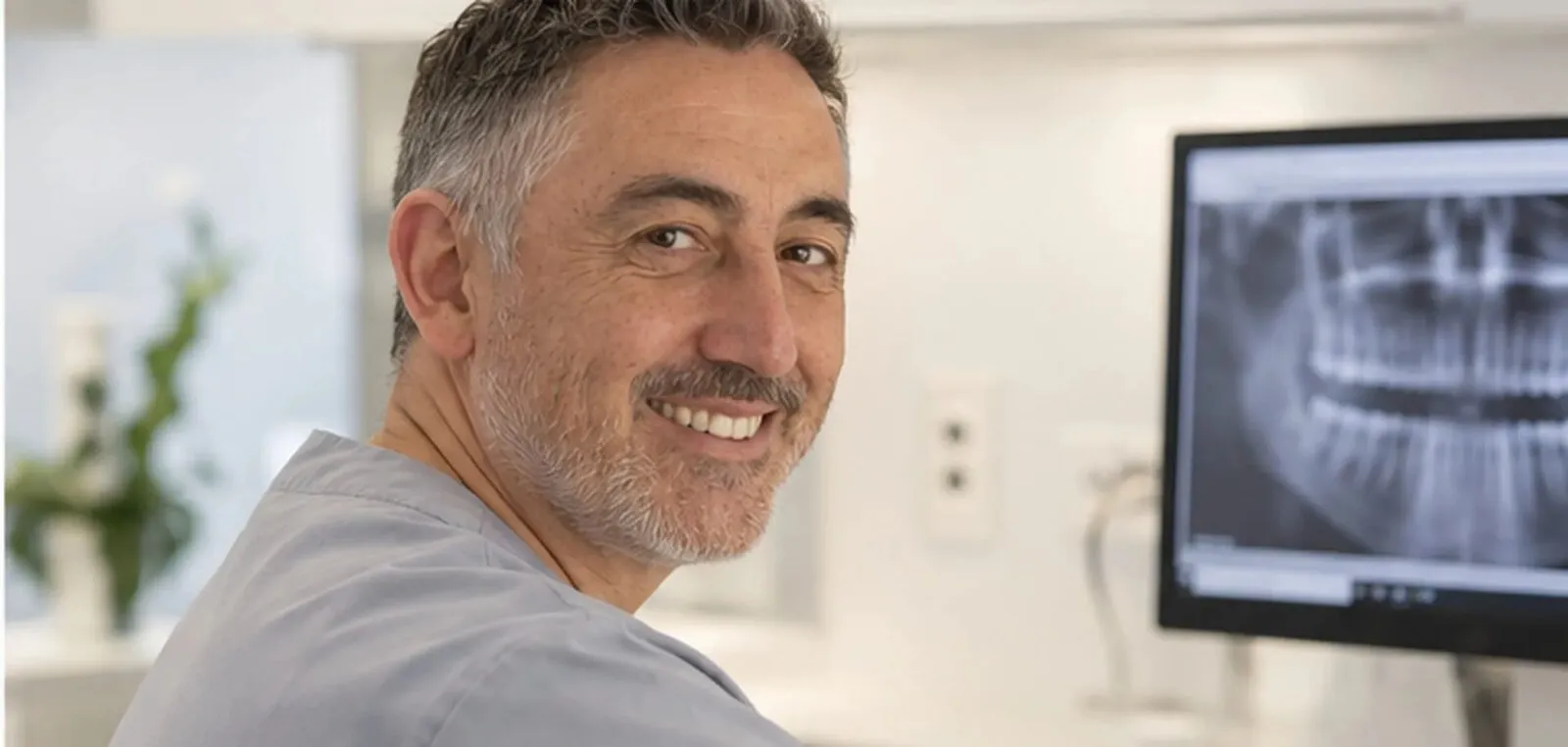 A dentist at his workstation, an X-ray of a full set of teeth on the monitor beside him
