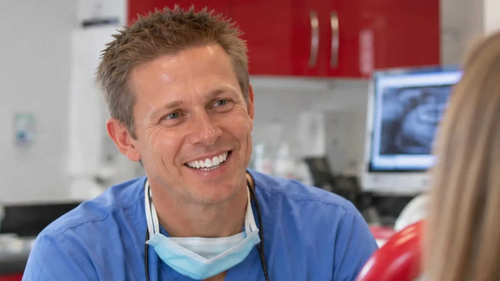 Dr Ross Cutts smiling in his surgery at Cirencester Dental Practice, red cabinets and an X-ray on the screen behind him