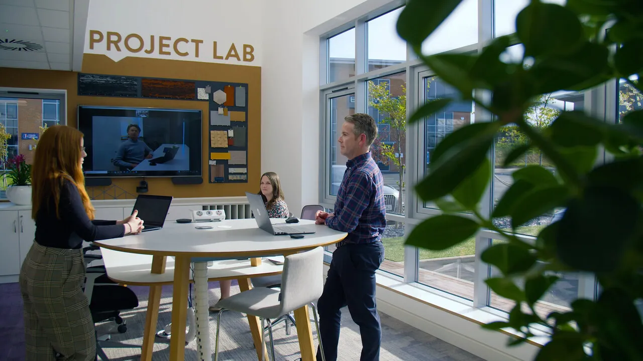 Two of the team in TecInteractive’s office, under a sign reading PROJECT LAB