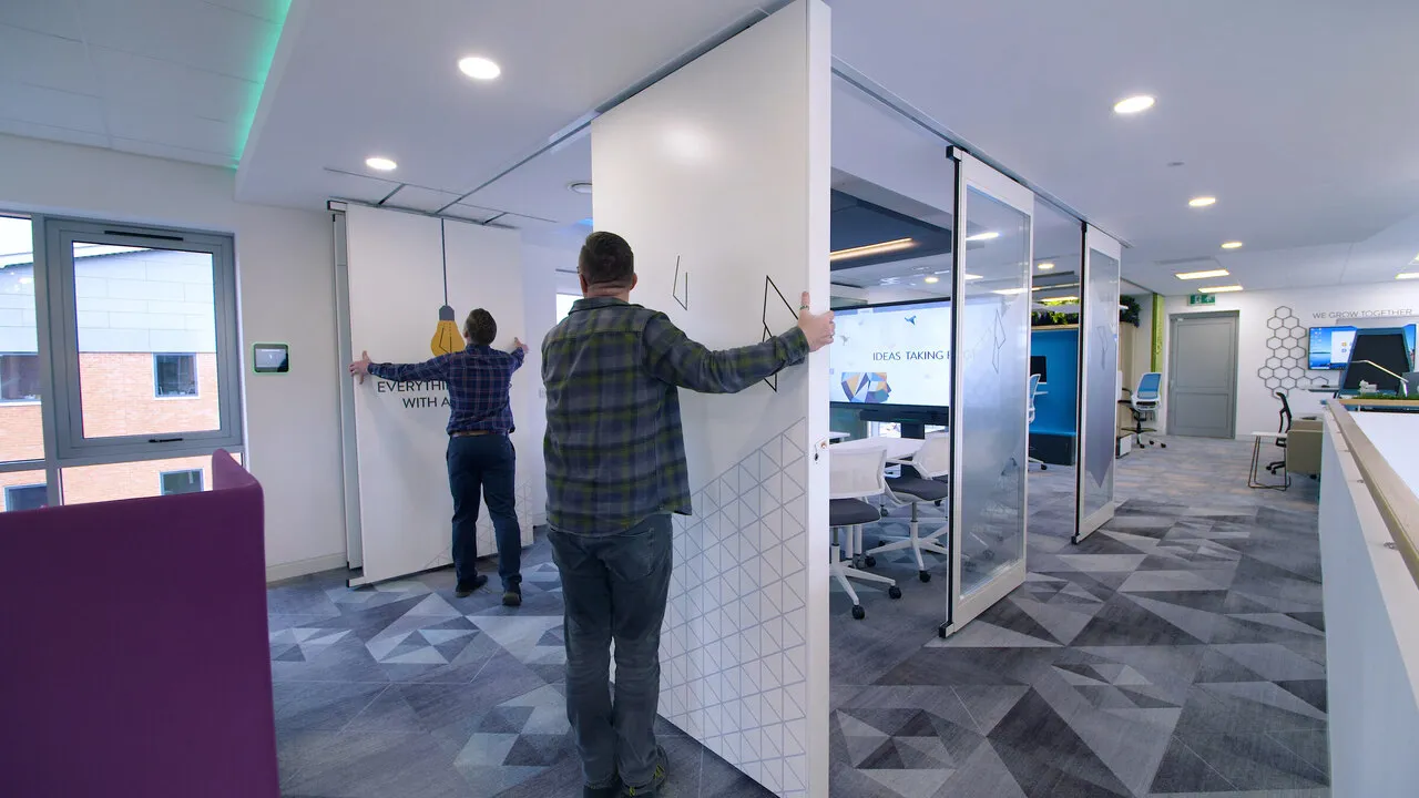 Two people drawing on a whiteboard wall in an open office