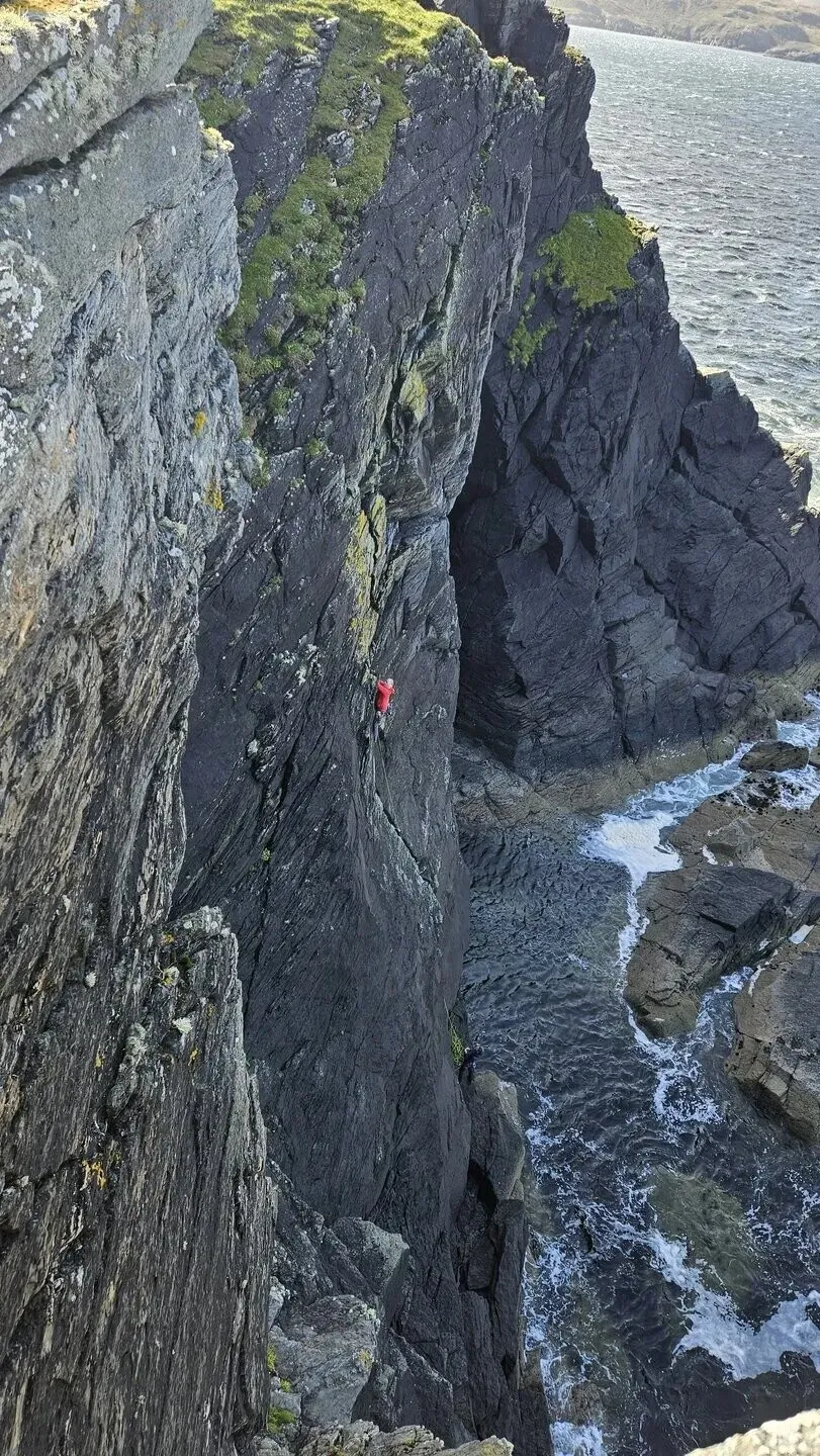 Looking down a dark sea cliff at Faraid Head: a climber in a red jacket halfway up a steep slab, the sea breaking on the rocks far below