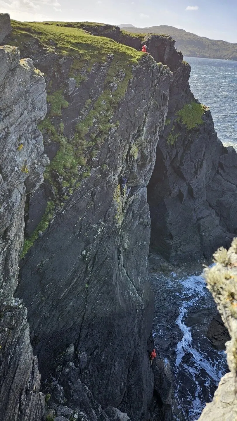 Three climbers on the cliff: one in red at the top, one halfway up the face and one at its foot by the sea