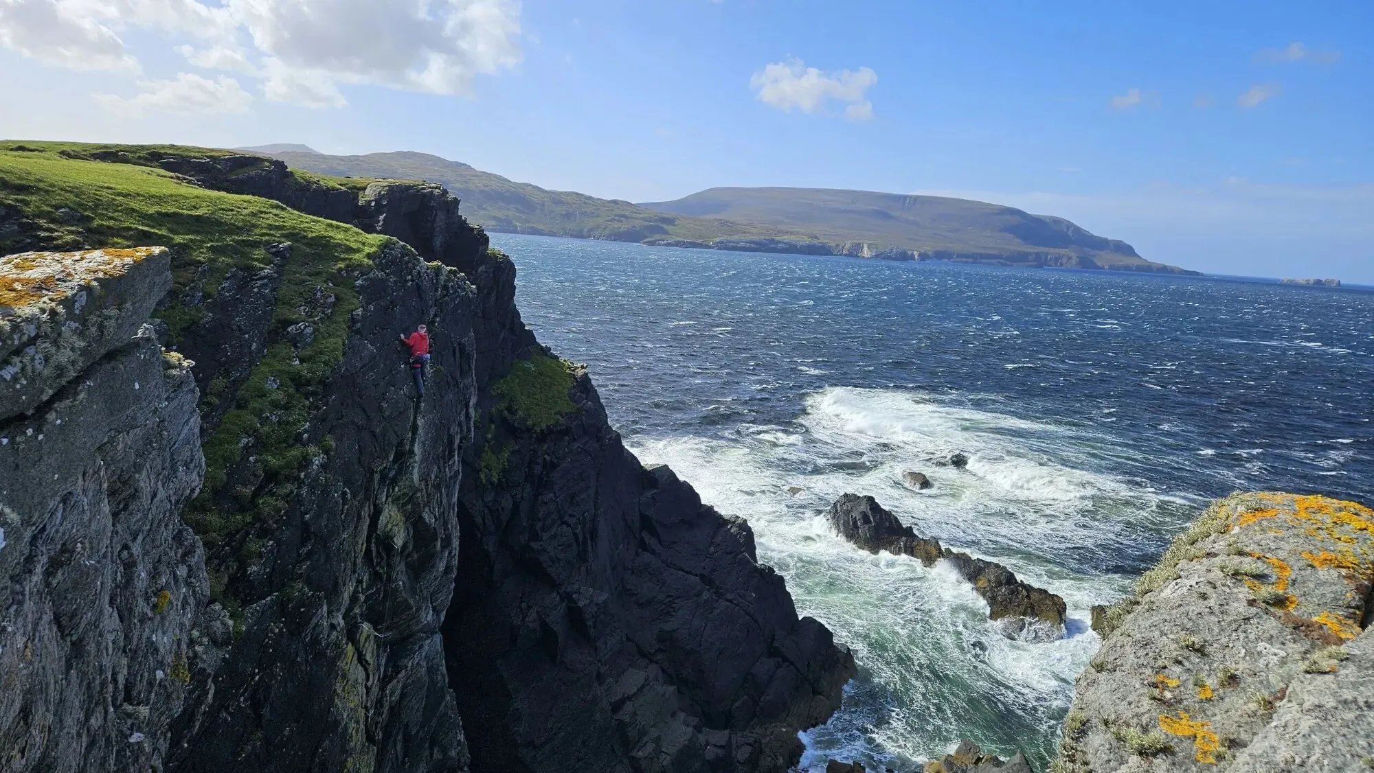 A climber in a red jacket on a sea cliff at Faraid Head above a rough blue sea, with the green headland and the hills of Sutherland beyond