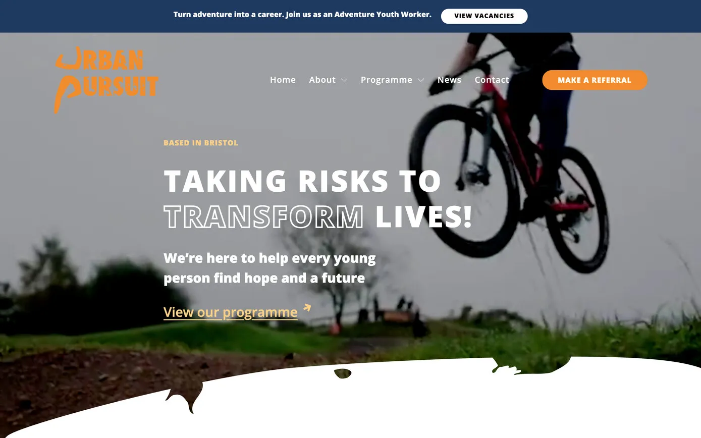 The Urban Pursuit homepage: the orange logo and menu over their film of a young rider mid-jump on a mountain bike, with “Taking risks to transform lives!” and a blue vacancies banner along the top