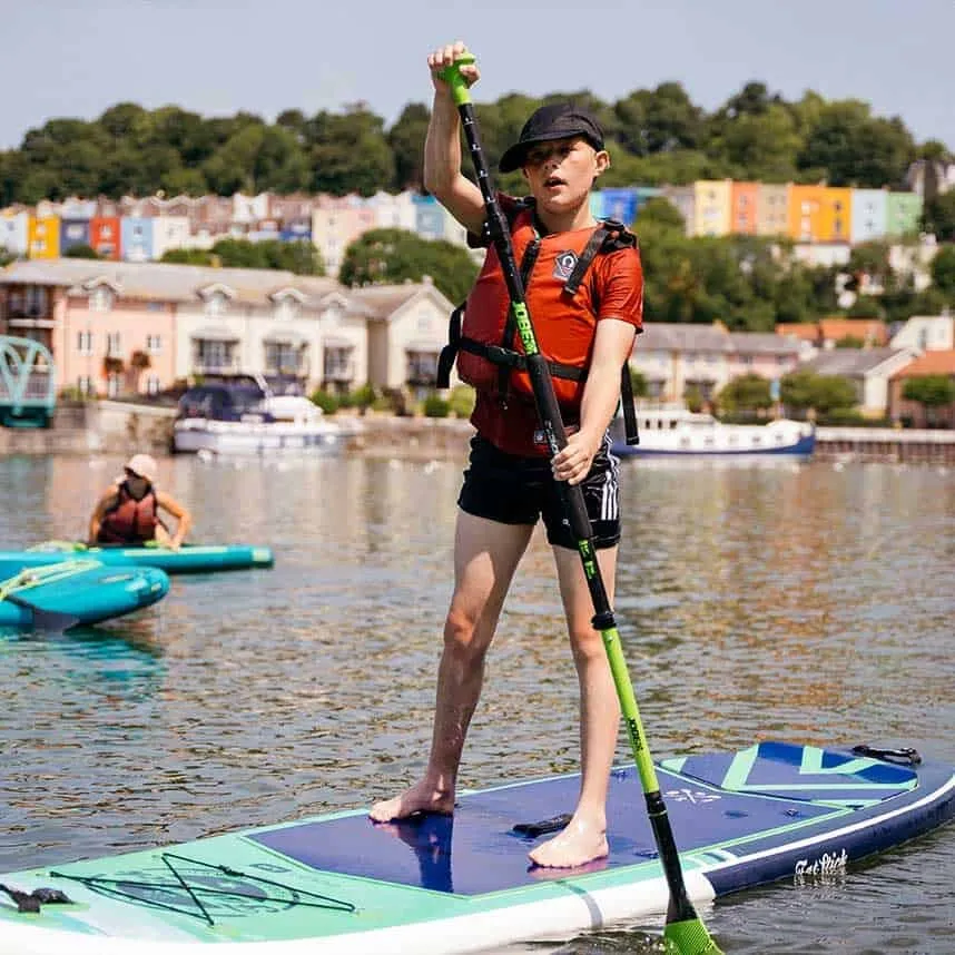 A young person on a paddleboard on Bristol’s harbour, one arm raised, with coloured houses behind