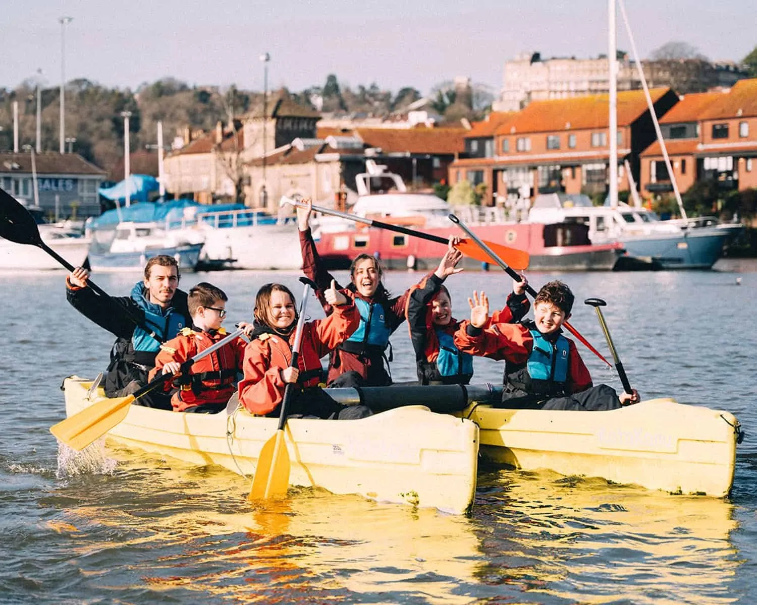 A group of young people cheering on rafts on Bristol’s harbour, paddles raised